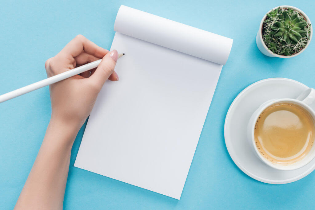 hand with pencil ready to write on blank piece of paper with plant and tea next to the paper on the right